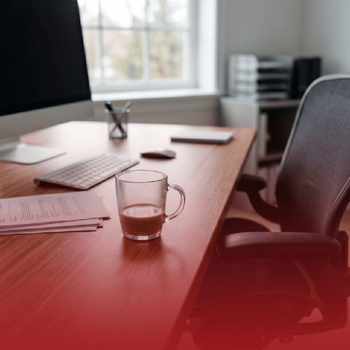 A bright, modern home office desk with a computer, keyboard, documents, and a glass mug of coffee. The bottom of the image features a transparent red color gradient.