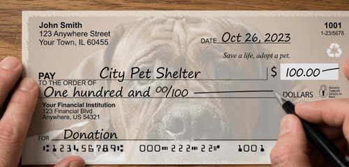 A person&rsquo;s hands writing a check for $100.00 to "City Pet Shelter." The check features a large, faded background image of a dog's face and includes the text "Save a life, adopt a pet.